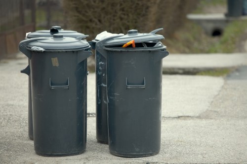 Two large, dark grey wheelie bins with hinged lids sit on a paved surface outdoors, likely on a driveway or street area. The bin on the left has a closed lid, while the one on the right has an open lid with some orange and white objects visible inside. Both bins are made of plastic with a matte finish and are positioned side by side, facing forward, with their handles and wheelsets partially visible at the base. In the background, blurred natural elements such as trees or bushes are visible, along with a curb and a glimpse of a bridge or tunnel in the distance, suggesting a residential or suburban environment. This scene aligns with rubbish collection services and waste management practices typical of the Holborn area, as handled by Garden Clearance Holborn, and visually emphasizes the need for proper waste disposal and clearance in a local context.