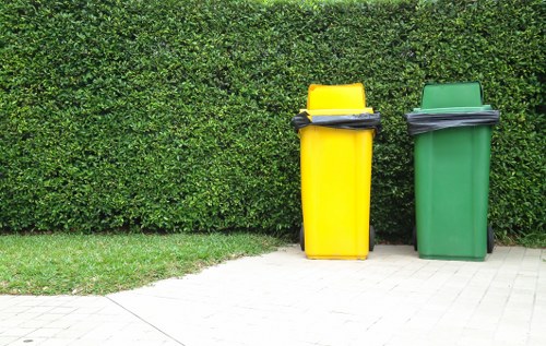 Two large wheelie bins, one yellow and one green, are positioned side by side on a paved area in front of a neatly trimmed hedge. The yellow bin, located on the left, has a closed lid with a black handle and appears to contain black rubbish bags or waste materials. The green bin, to the right, also has a closed lid with a black handle and seems to be used for general waste or recyclables. The paved surface consists of light-colored paving slabs, and the hedge behind the bins is dense and uniformly trimmed, providing a natural green backdrop. The overall scene appears to be outdoors in a residential or commercial garden area, typical for rubbish collection or waste removal services in the Holborn area. The image reflects the typical setup for rubbish disposal and aligns with garden clearance and waste removal activities offered by companies like Garden Clearance Holborn.