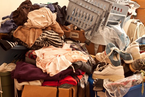 A large pile of assorted clothing, fabrics, and textiles including t-shirts, sweaters, and jackets in various colours and textures, with some items folded and others crumpled, situated on a cluttered surface. On top of the pile, there is a grey plastic laundry basket filled with more clothing and fabric items, leaning at an angle. Surrounding the pile are pairs of shoes, including sneakers and loafers, some placed randomly and others partially visible. The background features a domestic indoor setting, suggesting the clutter may be within a room or storage area. The image, associated with rubbish removal services in the Holborn area, highlights the need for efficient waste clearance of household textiles and clothing, typical of the kind of clutter handled by Garden Clearance Holborn near London postcode areas.