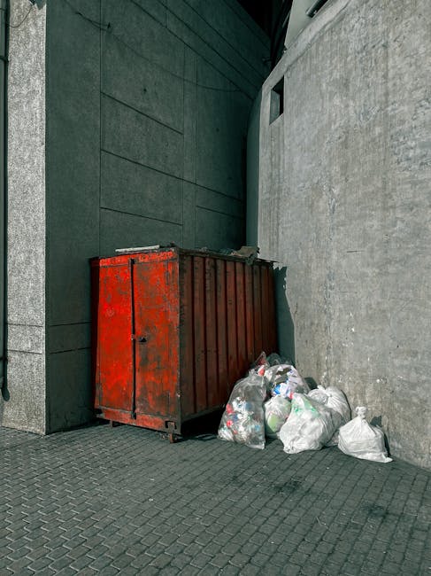 A disorganized collection of overflowing rubbish bins and scattered waste bags sits on the pavement in front of a commercial building, with various types of waste including cardboard boxes, paper, pla